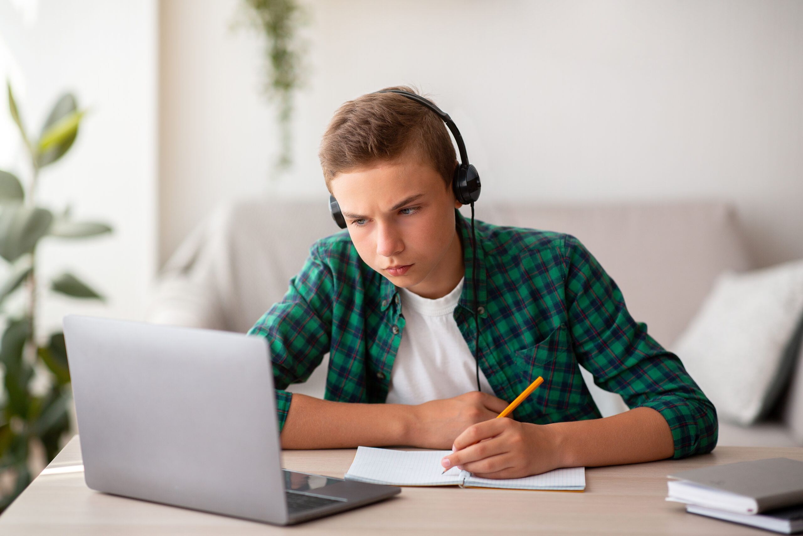 Concentrated school boy studying online from home, wearing headset, sitting in front of laptop and taking notes, copy space