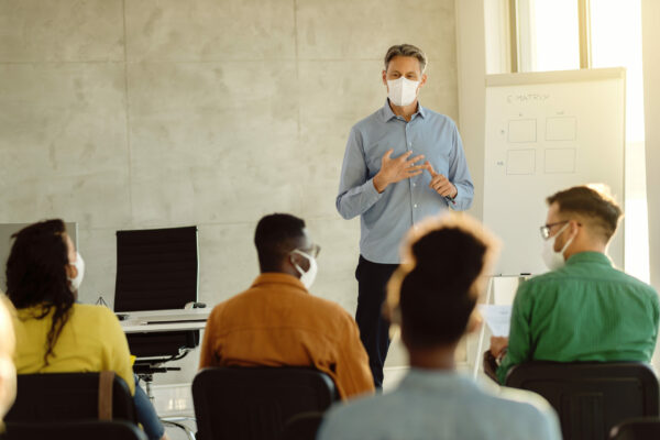 Mid adult teacher with protective face masks talking to a group of university students in lecture hall. Teacher wearing protective face mask while holding a class to group of teachers during coronavirus epidemic.