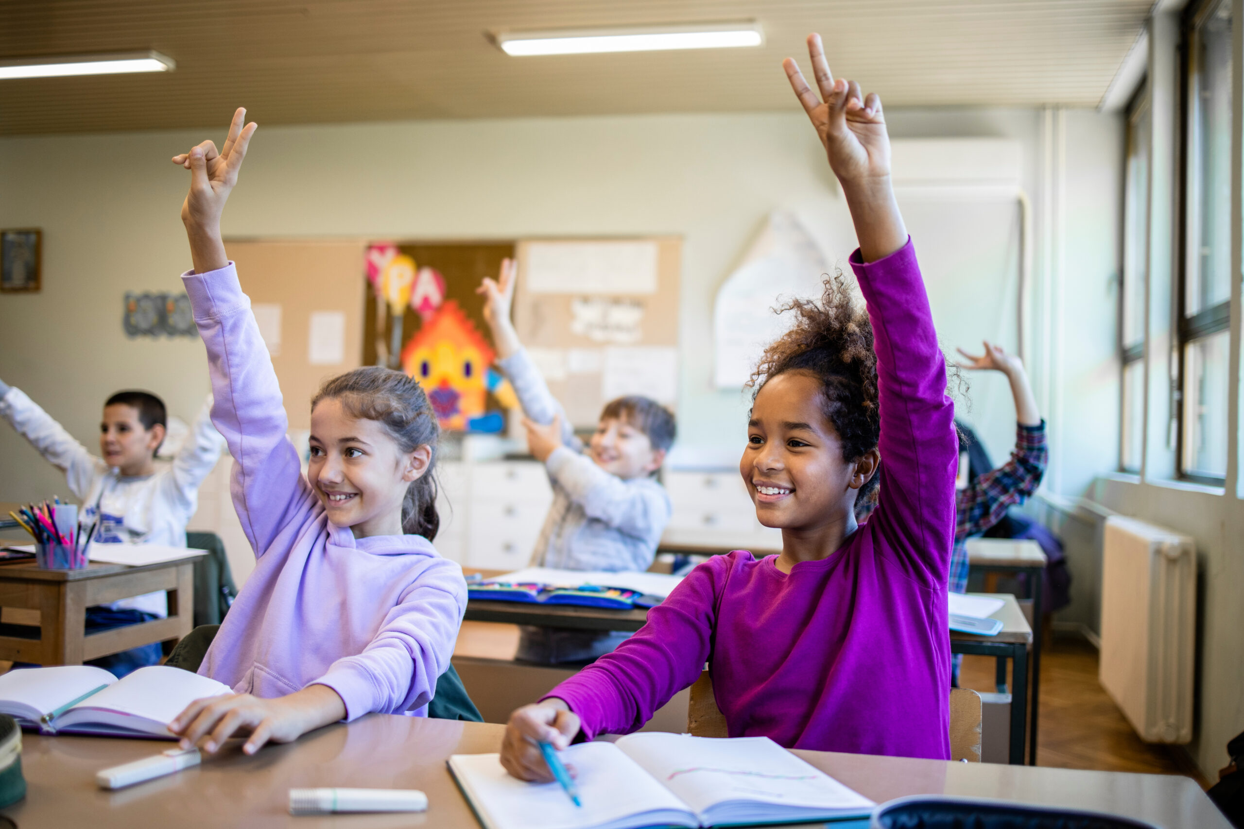 Schoolchildren at classroom with raised hands answering teacher's question and smiling.