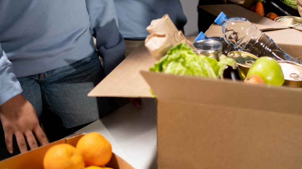 A cardboard box filled with food donations including fruit, bottled water and tin cans.
