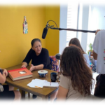 People talking at a table with a microphone, being filmed